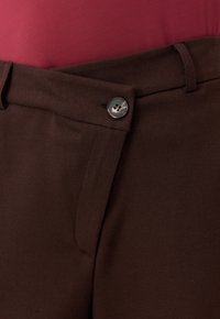 Zbliżenie na ciemnobrązowe spodnie z zapięciem w pasie i pętlą na pasek, noszone z czerwoną bluzką.