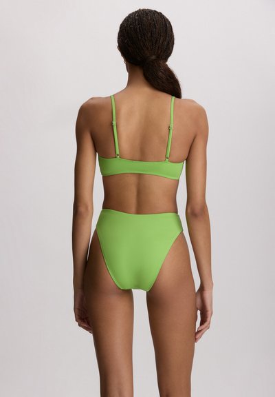Γυναίκα με πλεξούδες που φοράει μπικίνι φωτεινού πράσινου χρώματος, εμφανίζεται από πίσω πάνω σε ένα απλό ανοιχτό φόντο.