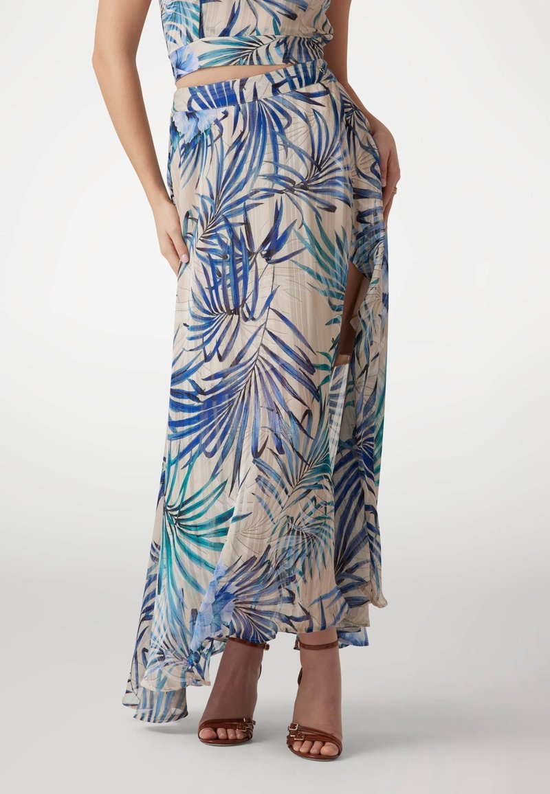 Falda maxi floral en tejido ligero y translúcido con patrones de hojas de palmera en azul y verde. Presenta una abertura alta y un dobladillo fluido. Sandalias de tacón marrón.