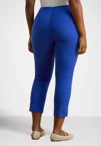 Nő áll, élénk kék, rövidített nadrágot és bézs hálós balettcipőt visel, hátulról látható, egyszínű fehér háttér előtt.