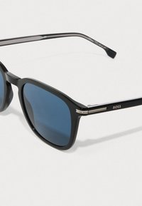 Μαύρα γυαλιά ηλίου με μπλε φακούς, ορθογώνιο πλαστικό σκελετό και μεταλλικά στοιχεία στους βραχίονες. Λογότυπο της μάρκας σε έναν βραχίονα.