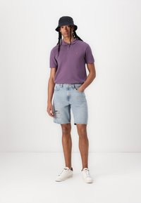 Polo shirt viola a maniche corte, pantaloni shorts in denim azzurro chiaro strappati, sneakers bianche e un cappello a secchiello nero, in piedi su uno sfondo neutro.