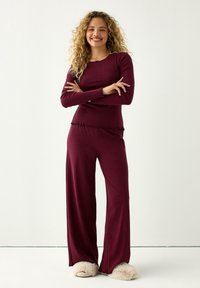 Burgundy lounge-set med långa ärmar och vida byxor, texturerad tyg och dekorerad söm vid fållen. Matchas med fluffiga tofflor.
