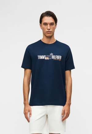 Άνδρας που φοράει μπλε σκούρο t-shirt Tommy Hilfiger με γραφικό παραλίας και άσπρο σορτς, στέκεται μπροστά σε απλό ανοιχτό φόντο.