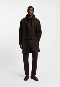 Cappotto marrone scuro, felpa coordinata e pantaloni su misura. Tessuto liscio con un design strutturato. Scarpe in pelle marrone completano il look.