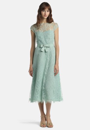 Žena v svetlomodrých midi šatách z krajky so sieťovanými krátkymi rukávmi a opaskom s mašľou, v kombinácii so zlatými sandálmi s otvorenou špičkou.