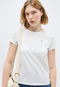 Νεαρή γυναίκα με κοντά ανοιχτό καστανά μαλλιά φοράει ένα απλό λευκό t-shirt και κρατάει μια εκρού τσάντα ώμου με χρυσή αγκράφα.