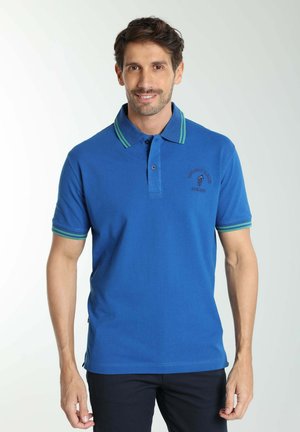 Homme souriant portant un polo bleu avec un col et des manches rayés verts, debout devant un fond uni clair.