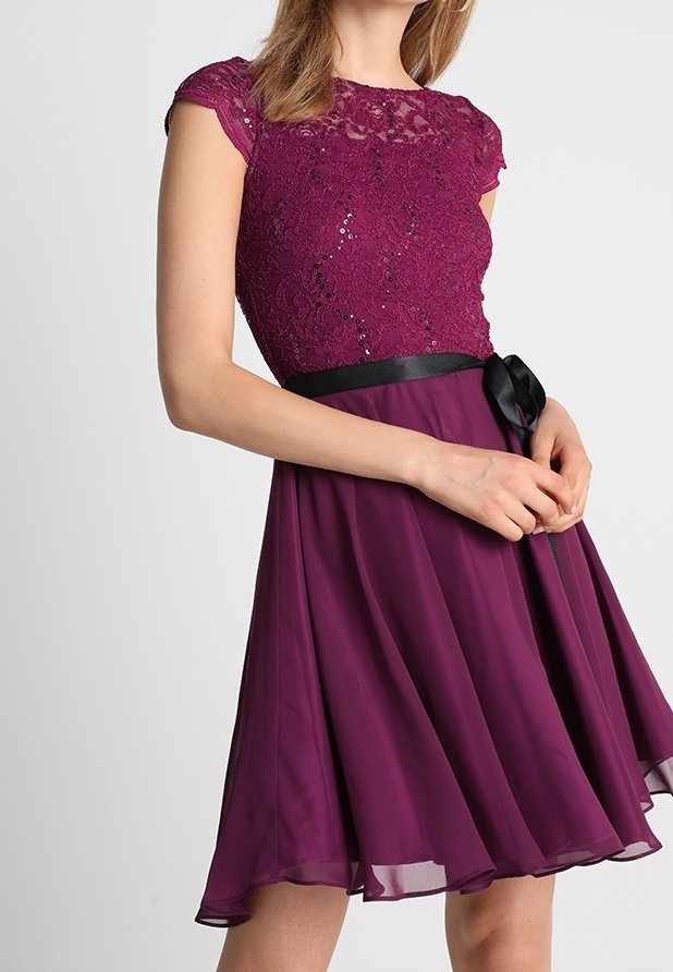 Robe prune sans manches avec corsage en dentelle pailleté, ceinture ruban noire et jupe fluide en chiffon, portée par une femme à la peau claire et aux cheveux blonds.