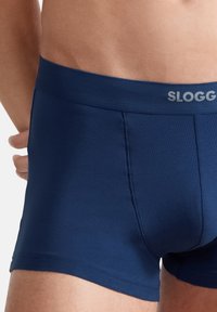 Marineblaue Boxer-Shorts aus glattem Stoff, mit einem weichen elastischen Bund, auf dem der Markenname "SLOGG" in Hellgrau aufgedruckt ist.