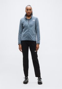 Μπλε φλις μπουφάν με φερμουάρ, συνδυασμένο με μαύρο παντελόνι και μαύρα παπούτσια. Το μπουφάν διαθέτει ψηλό γιακά και λογότυπο στο στήθος.