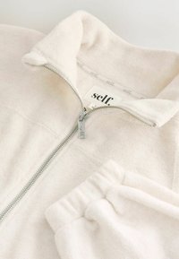 Weiße Fleece-Reißverschlusjacke mit hohem Kragen und einer Seitentasche. Enthält einen silbernen Reißverschluss und ein Markenetikett im Kragen.