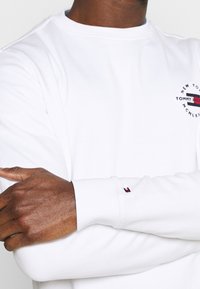 Felpa bianca con polsini a costine, caratterizzata da un logo ricamato circolare con accenti rossi, bianchi e blu. Tessuto liscio, vestibilità comoda.