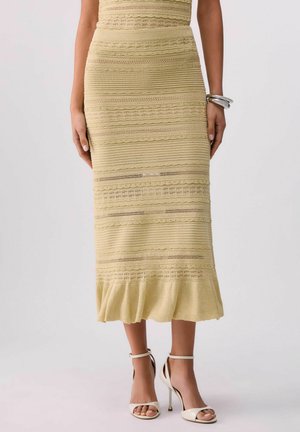 Donna che indossa una gonna midi gialla con texture e orlo arricciato, sandali bianchi con tacco alto e braccialetti d'argento a un polso.