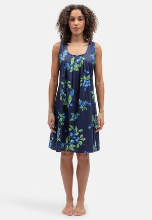 Frau steht barfuß und trägt ein ärmelloses marineblaues Kleid mit grünem Blatt- und blauem Beerenmuster.