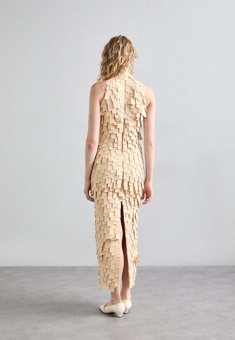 Beige, sleeveless dress featuring layered square pieces, textured with a long back slit, paired with light-colored shoes.