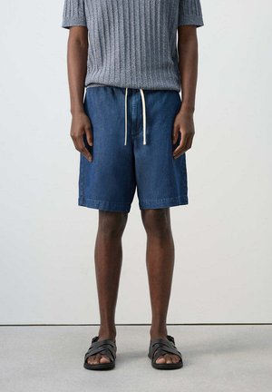 Persona con pantalones cortos vaqueros azules con cordón blanco, camiseta de punto gris de manga corta y sandalias negras con tiras cruzadas, de pie contra una pared lisa.