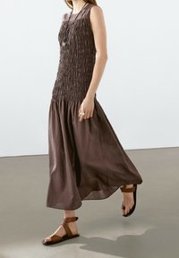 Brun, ärmlös, smockad klänning med en flödande kjol. Tyget är strukturerat; matchas med bruna sandaler med remmar och en ring.