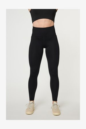 Žena stoji noseći crni sportski grudnjak, crne tajice visokog struka i bež tenisice na jednobojnoj svijetloj pozadini.