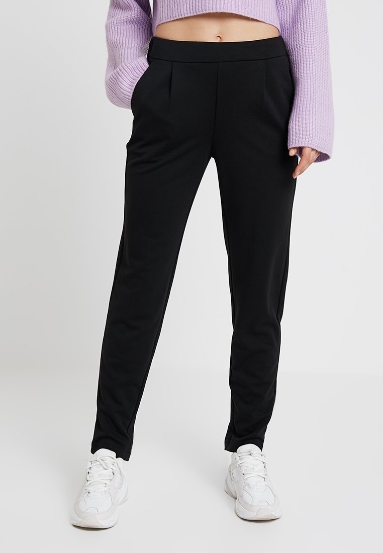 Personne portant un pantalon noir fuselé, des baskets blanches et un pull court lavande à manches longues, debout devant un fond uni.