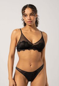 Zwarte kanten bralette met een doorschijnend ontwerp, voorzien van verstelbare bandjes en gegolfde randen. Gecombineerd met bijpassende doorzichtige zwarte slipjes.