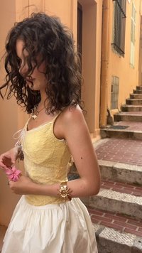 Yellow lace top with thin straps, paired with a white skirt. Accessories include a gold bracelet and a pink flower in hand. Rustic background.