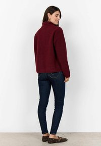 Burgundy színű, textúrázott pulóver magas gallérral, sötét, skinny farmernadrággal és barna lapos cipővel párosítva, hátulról nézve.