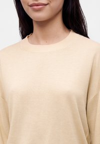 Femme portant un pull beige à col rond, montrant le bas du visage, le cou et le haut de la poitrine avec de longs cheveux foncés visibles.