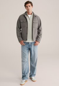 Grijze wollen blend jas met twee borstzakken, gelaagd over een lichtgroene zip-up trui, gecombineerd met lichtblauwe jeans en witte sneakers.