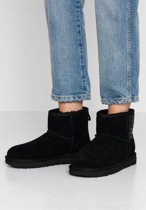 Jambes portant un jean bleu clair et des bottines en daim noir doublées de fourrure, debout sur un sol blanc lisse.