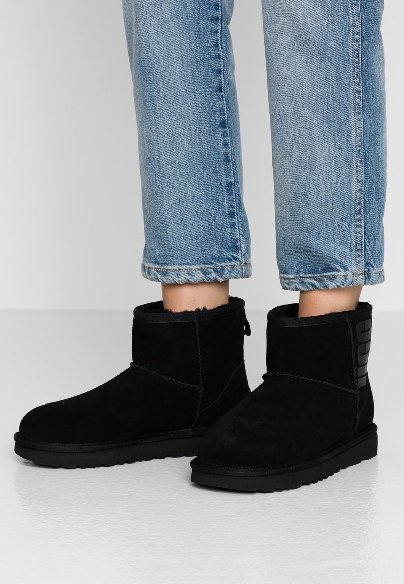 Jambes portant un jean bleu clair et des bottines en daim noir doublées de fourrure, debout sur un sol blanc lisse.