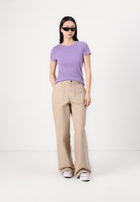 T-shirt rigato lilla chiaro e pantaloni beige a gamba larga, abbinati a occhiali da sole neri e sneakers bianche. Design minimalista, vestibilità casual.