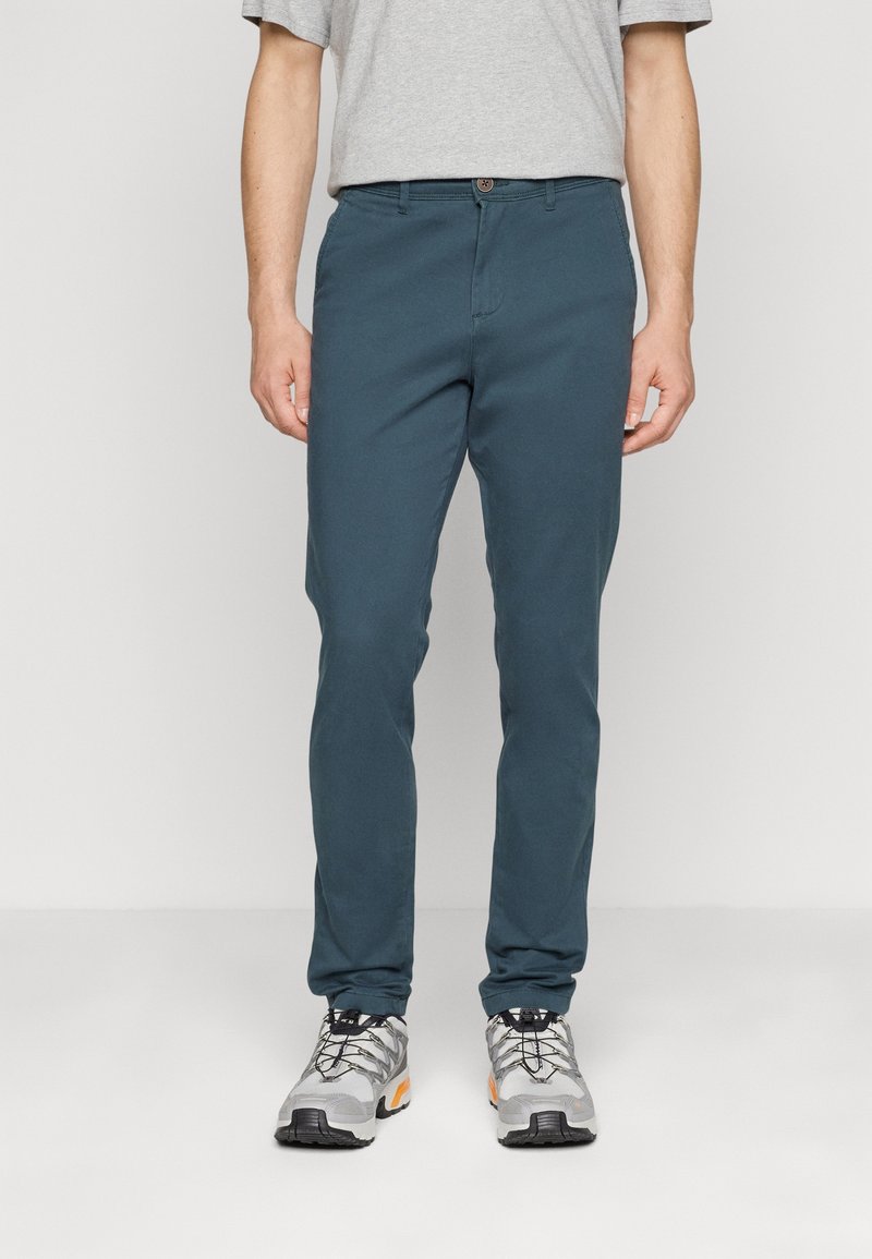 Homme portant un pantalon slim bleu sarcelle, des baskets grises à lacets noirs et une chemise à manches courtes gris clair, debout devant un fond uni.