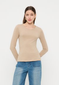 Γυναίκα με καστανά ίσια μαλλιά που φορά χρυσούς κρίκους στα αυτιά, μπεζ μακρυμάνικη μπλούζα και μπλε τζιν, σε λευκό φόντο.