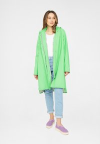 Chubasquero impermeable verde brillante, de longitud media y con capucha, cierre delantero de botones a presión, combinado con unos vaqueros azul claros y zapatos morados tipo slip-on.