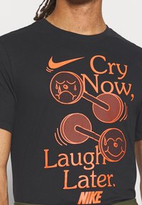 Camiseta de algodón negra con texto impreso en naranja: "Llora ahora, ríe después" con ilustraciones de mancuernas y caras expresivas.