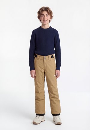 Lächelnder Junge mit navyblauem Pullover, beigen Schneehosen und weißen Winterstiefeln steht vor einem einfarbigen weißen Hintergrund.