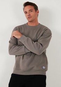 Grijze sweatshirt met een ronde halslijn, ribgebreide manchetten en zoom. Bevat een klein logo-op een patch op de onderzijde rechts. Gladde textuur.