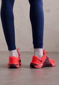 Nike sneakers in bright pink with black accents, featuring a textured design and triangular pattern on the sides. Worn with blue leggings.