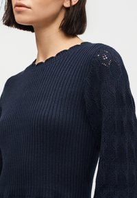 Γυναίκα που φοράει μπλε σκούρο πουλόβερ με ριμπ ύφανση, με μανίκια από δαντέλα με σχέδια και λαιμόκοψη με κυματιστό τελείωμα, εμφανίζεται από τους ώμους μέχρι το πηγούνι.