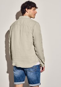 Světlá šedá lněná košile s dlouhými rukávy a detailem sklady na zadní straně, nošena s modrými džínovými šortkami. Pozadí je neutrální, což zvýrazňuje outfit.