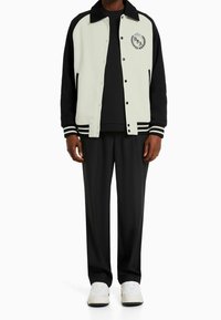 Svart och krämfärgad varsityjacka med svart krage, randiga detaljer och logotyp, i kombination med svarta byxor och vita sneakers.