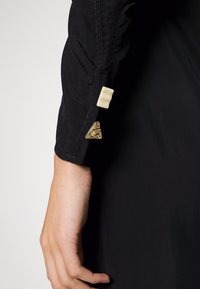 Svart tygärm med en slät textur. Har en vit rektangulär knapp och en guldfärgad triangulär accent nära manschetten.