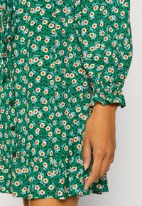 Groene bloemenjurk met oranje bloemen en zwarte stippen, knoopdetails en gathered mouwen met een gerimpelde zoom.