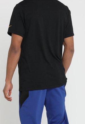 Personne portant un t-shirt noir à manches courtes et un pantalon de sport bleu, debout dos à la caméra.