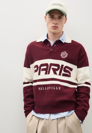 Junger Mann trägt einen bordeauxroten und weißen Pullover mit dem Text "PARIS BELLEVILLE", beige Hose, blau gestreiftes Hemd und eine weiße Baseballkappe.