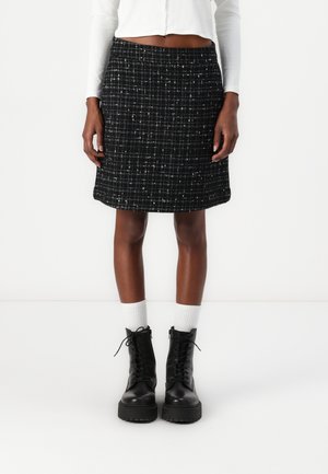 Jupe en tweed noir avec un motif texturé, coupe droite, mi-longue, associée à des bottes noires à lacets et des chaussettes blanches taille bateau.
