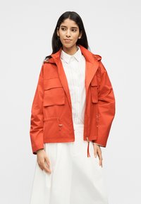 Femme portant une veste orange vif avec de grandes poches avant par-dessus une chemise blanche à col et une jupe blanche fluide, debout avec les mains détendues.