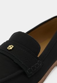 Mocasinii negri din piele au un finisaj texturat, un accent pătrat pe bareta și o talpă maro. Designul include detalii subtile de cusătură.