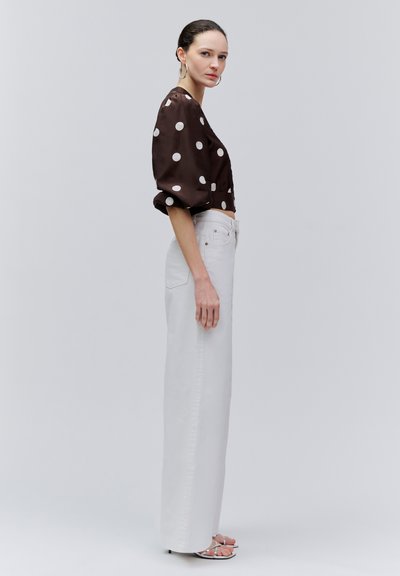 Naine, kellel on taha kammitud juuksed, hõbedased rõngaskõrvarõngad, pruun täpiline pluus, kõrge vöökohaga valged laiade pükste jalaosadega püksid ja hõbedased kontsakingad, seisab külje pealt.